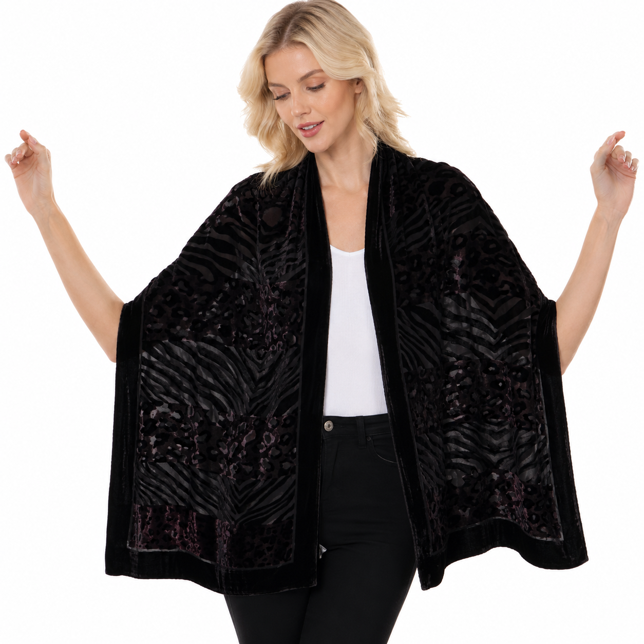 A woman modeling a black open-front shawl with a textured zebra-like pattern and velvet trim, worn over a white top and black pants against a plain white background.